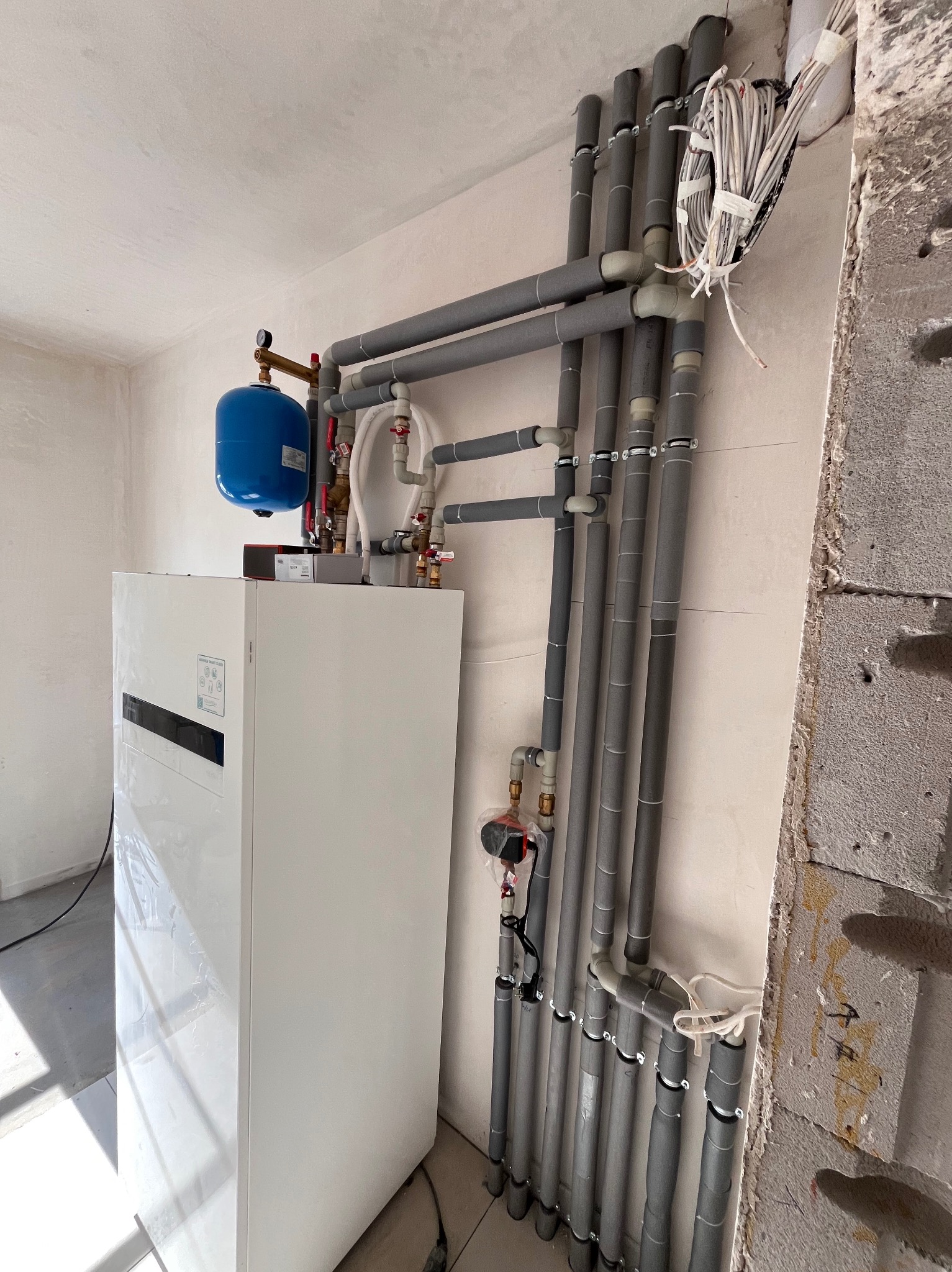 Biała pompa ciepła z niebieskim zbiornikiem i szarymi rurami w Sochaczewie. Instalacja w surowym wnętrzu, widoczne przewody i fragment ściany z betonu komórkowego.