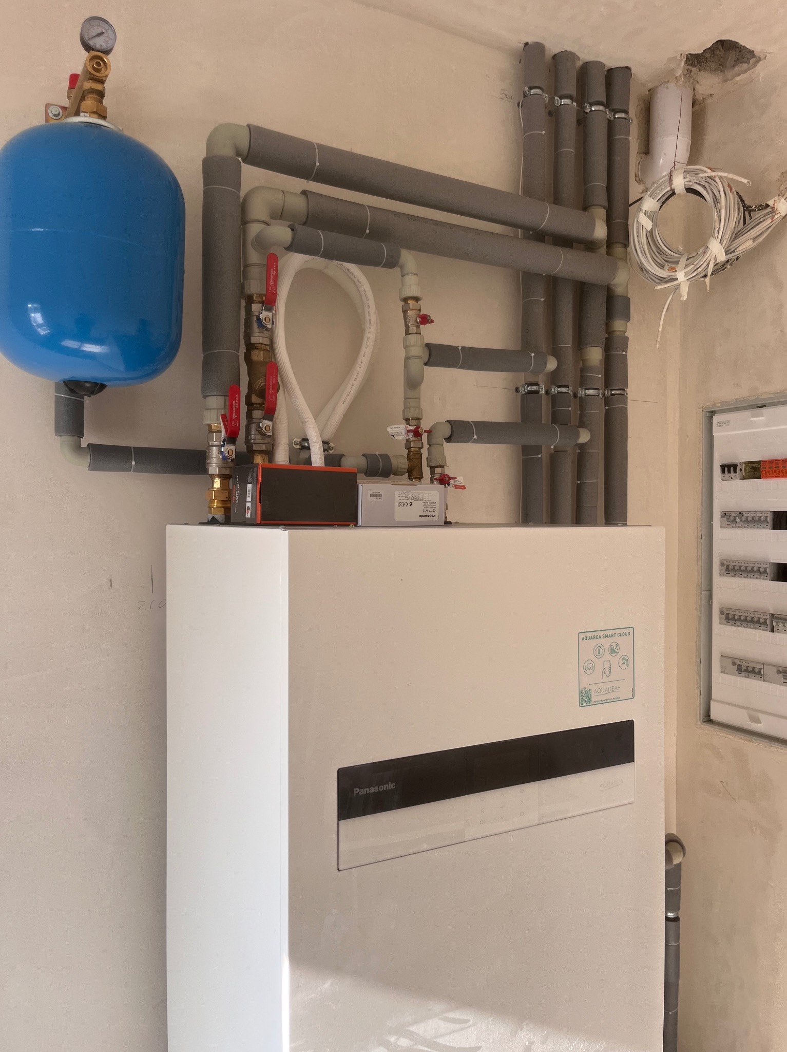 Biała pompa ciepła Panasonic z niebieskim zbiornikiem wyrównawczym i szarymi rurami w surowym wnętrzu. Widoczna instalacja elektryczna i zawory.