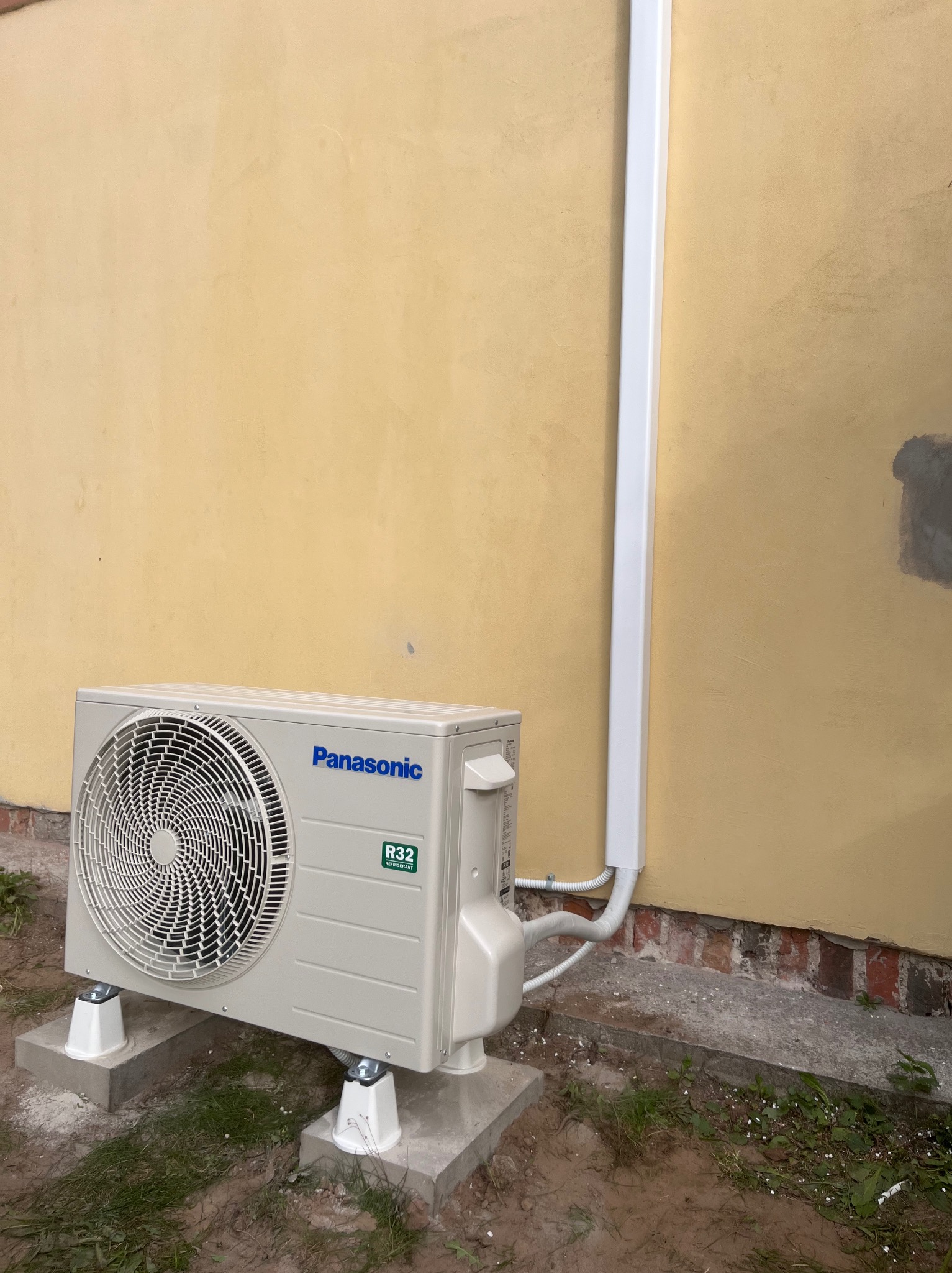 Zamontowana jednostka zewnętrzna klimatyzacji Panasonic R32 na betonowych podstawach przy żółtej ścianie budynku z białą rurą odprowadzającą. Widok z dołu.