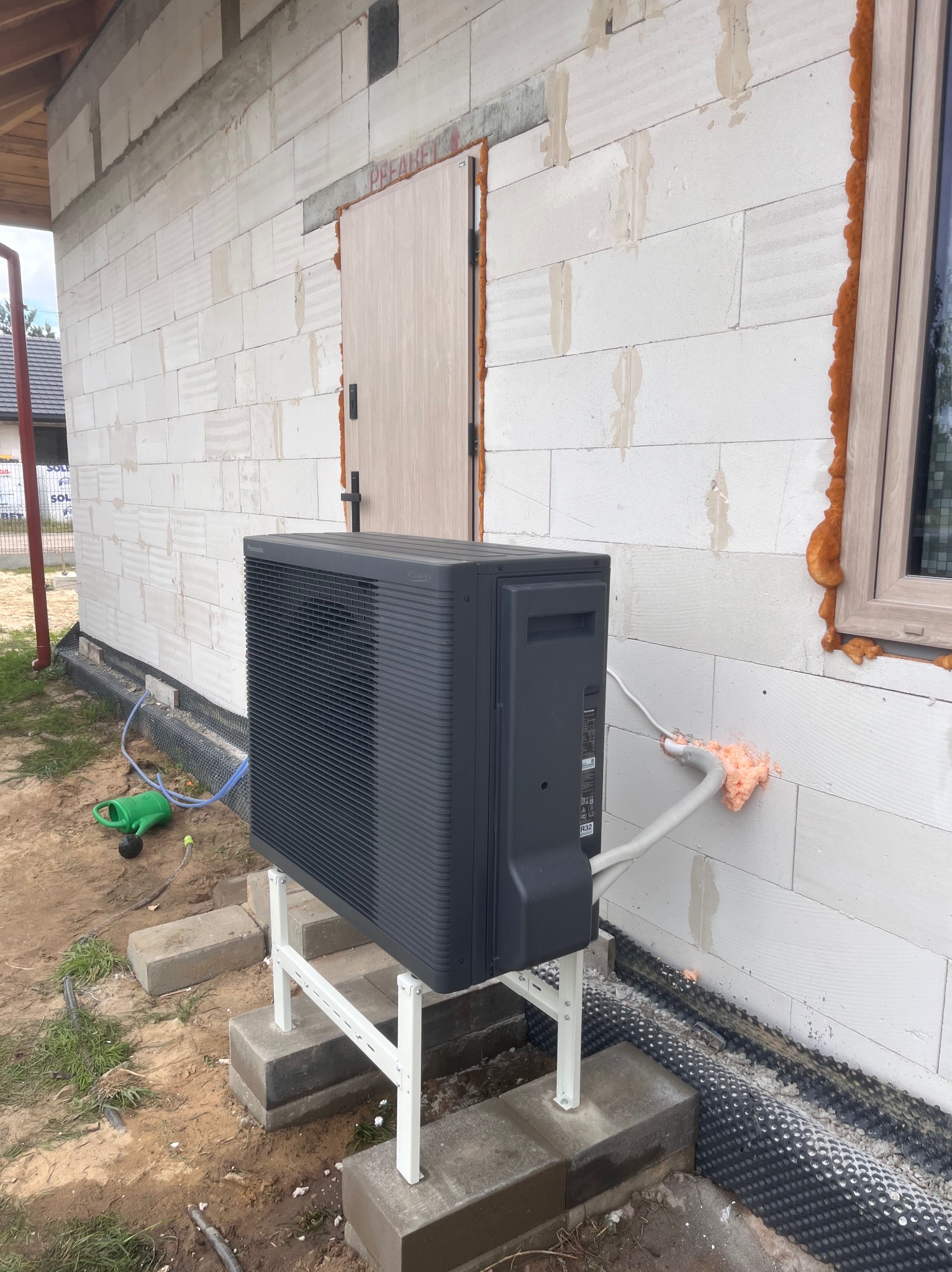 Szara pompa ciepła Panasonic Aquarea zamontowana na zewnątrz budynku z białego bloczka, z widocznymi elementami instalacji i ocieplenia okna pianką.