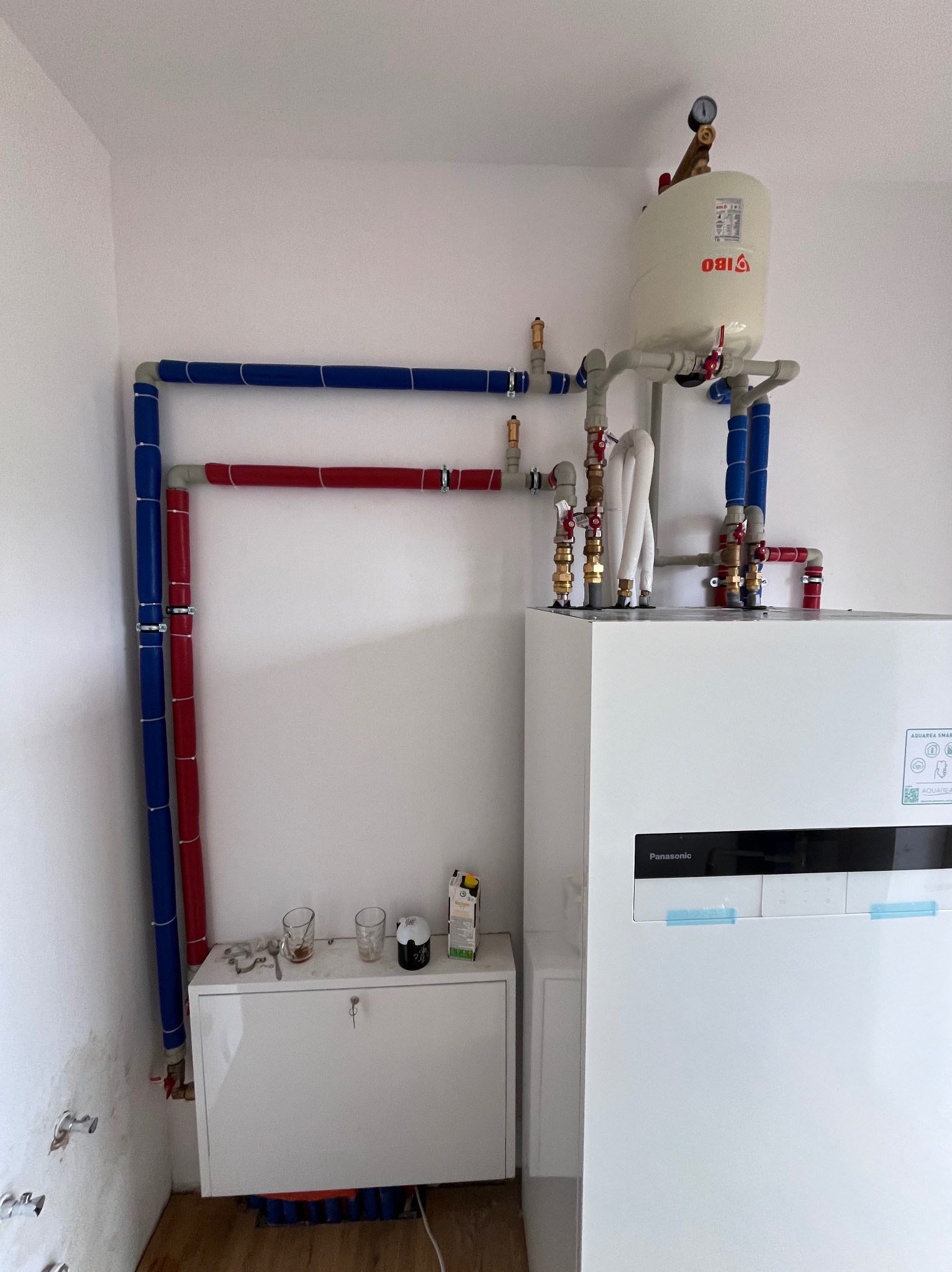 Biała pompa ciepła Panasonic z zbiornikiem IBO, połączona rurami w kolorach niebieskim i czerwonym. Widoczne elementy instalacji hydraulicznej i manometr.