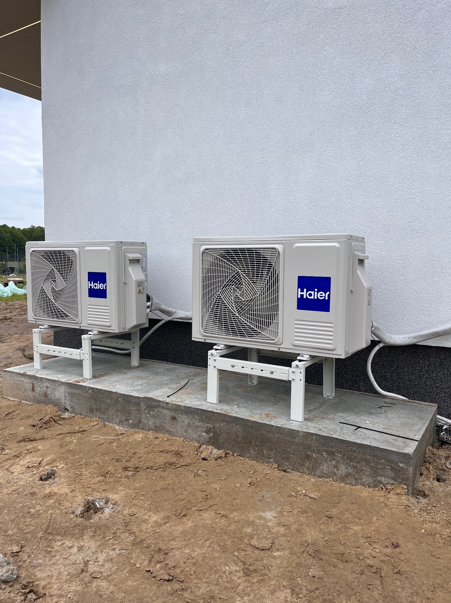 Dwa klimatyzatory Haier zamontowane na białych wspornikach przy ścianie budynku z widocznymi przewodami. Ujęcie z poziomu gruntu, podkreślające instalację.