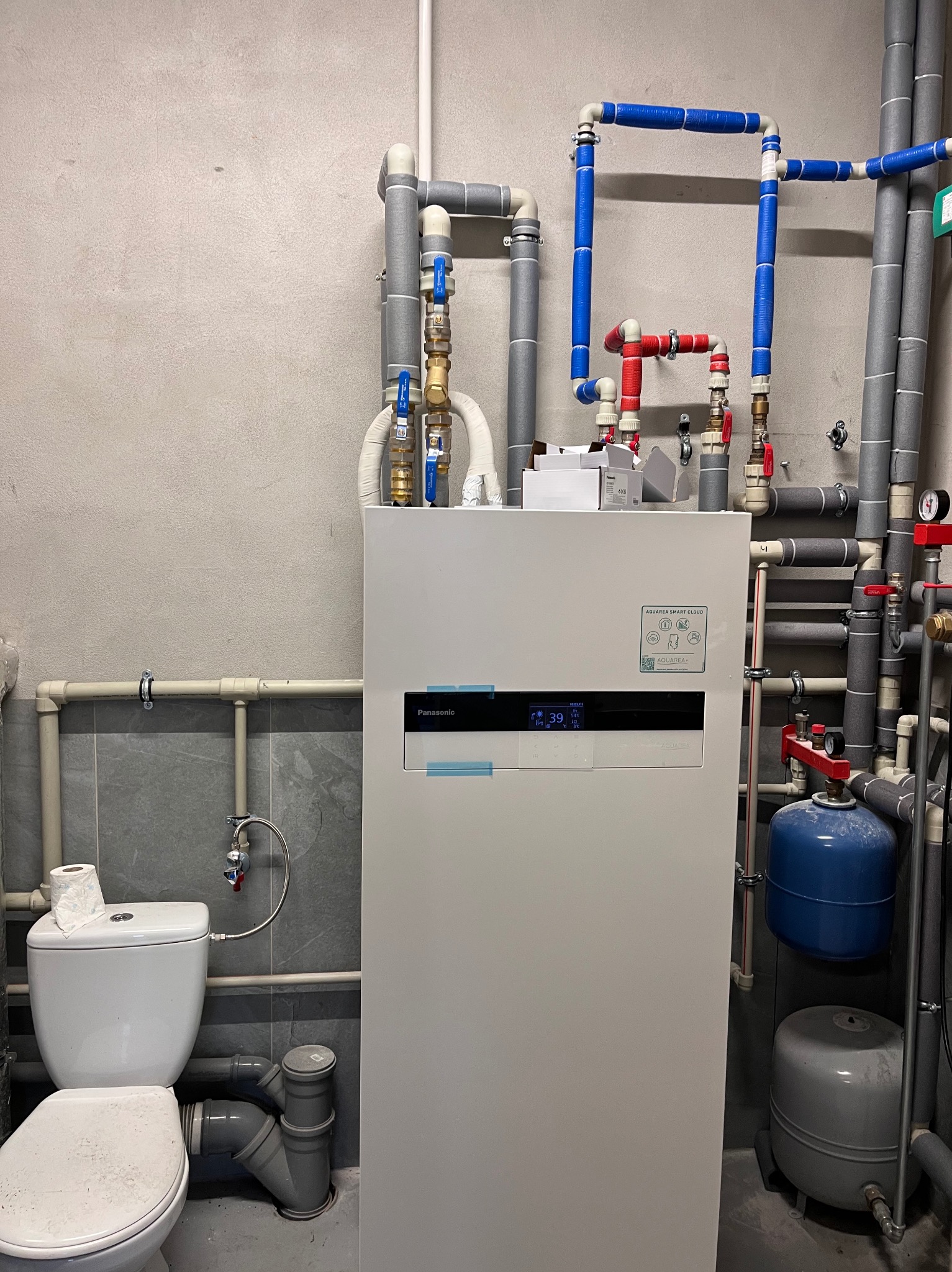 Biała pompa ciepła Panasonic Aquarea z licznymi rurami (szare, niebieskie, czerwone) w pomieszczeniu technicznym. Widoczny fragment toalety. Szare ściany.