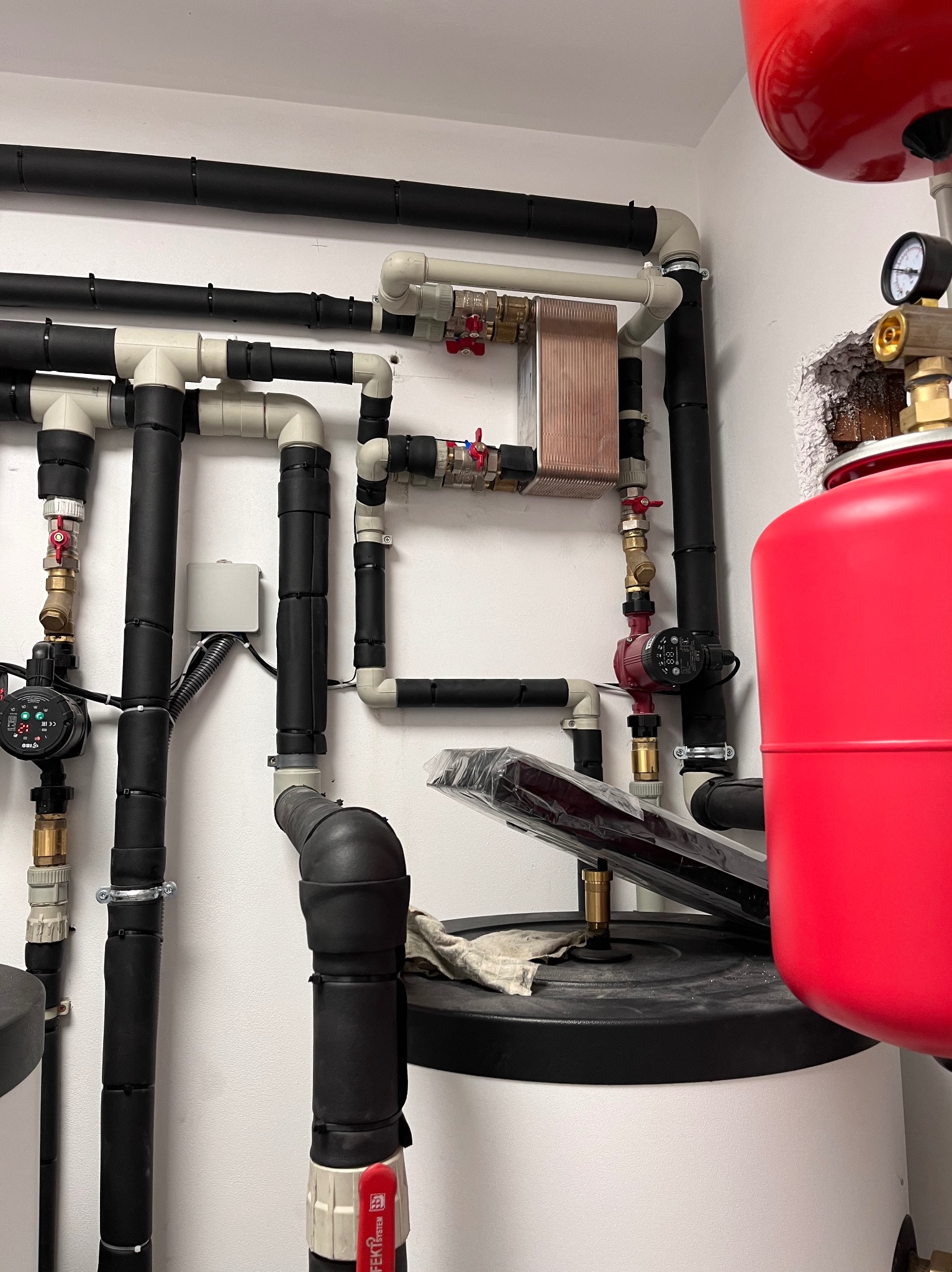 Złożona instalacja grzewcza z rurami w izolacji, miedzianym wymiennikiem ciepła i czerwonym zbiornikiem wyrównawczym na tle białej ściany. Widoczne zawory i pompy obiegowe.