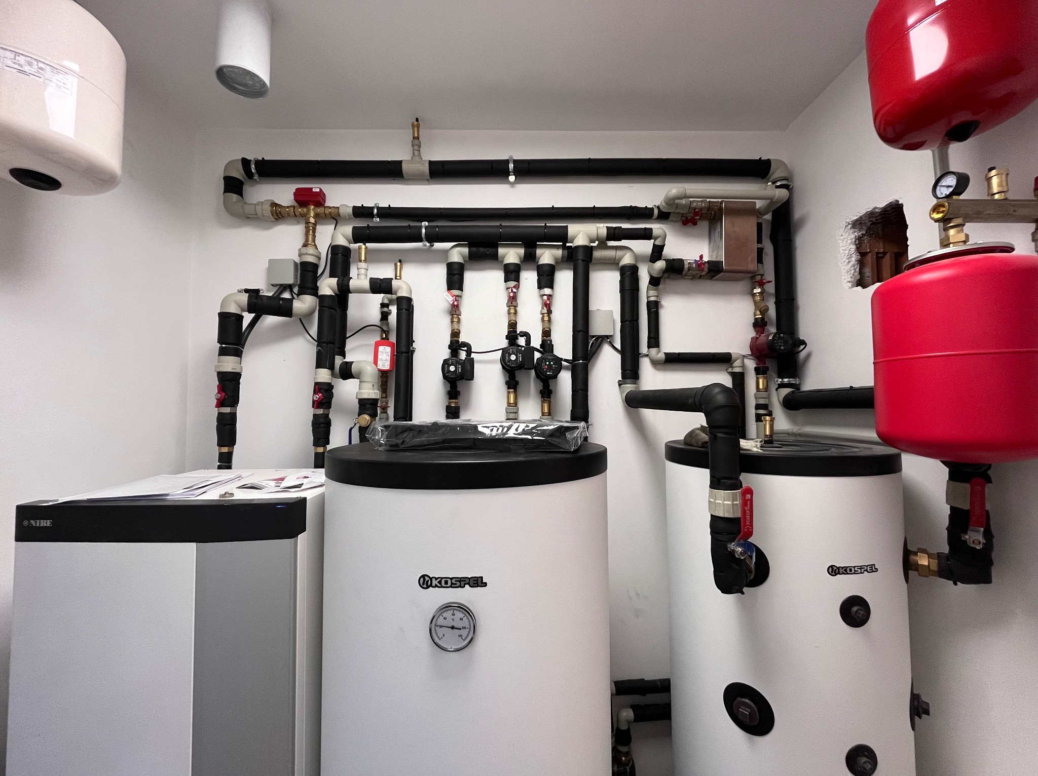Złożona instalacja grzewcza z dwoma zbiornikami Kospel i piecem Nibe, rury w izolacji, liczne zawory i manometry na białym tle ściany.