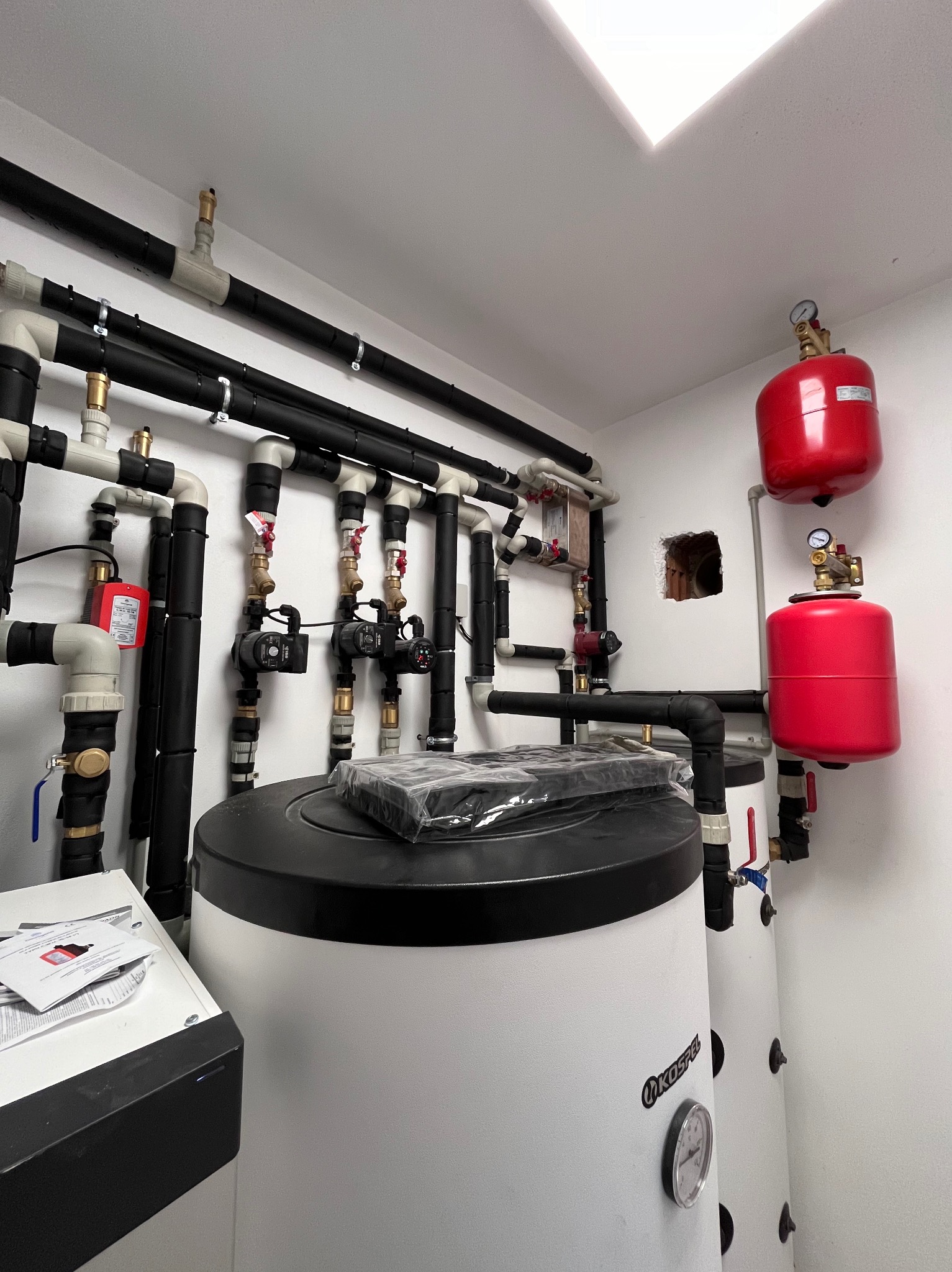 Biały zasobnik ciepłej wody użytkowej Kospel z czarną pokrywą, licznymi rurami i czerwonymi zbiornikami wyrównawczymi na białej ścianie w kotłowni.