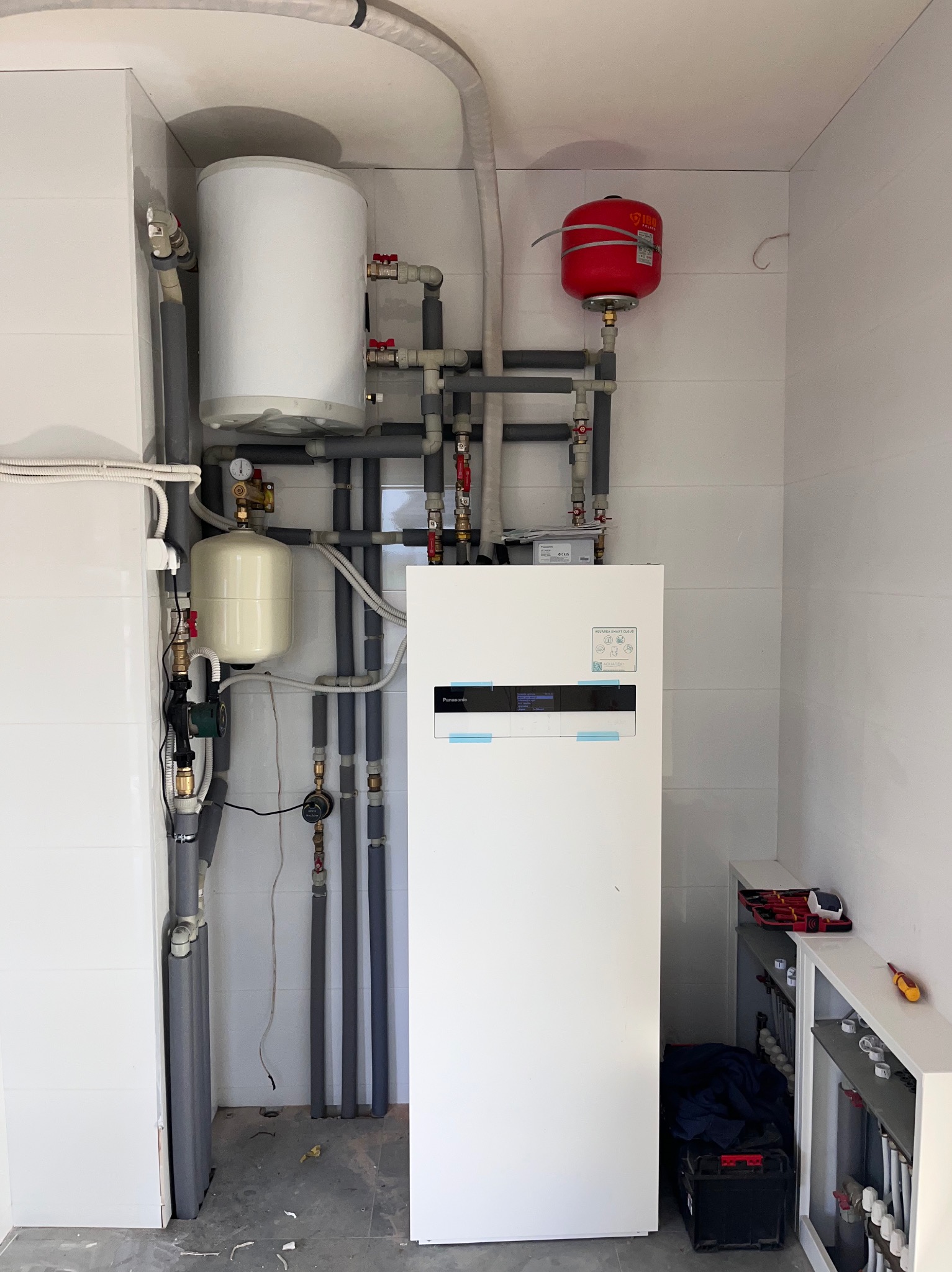 Biała pompa ciepła Panasonic z podłączonymi rurami i zbiornikami w pomieszczeniu z białymi płytkami. Widoczne narzędzia na półce.