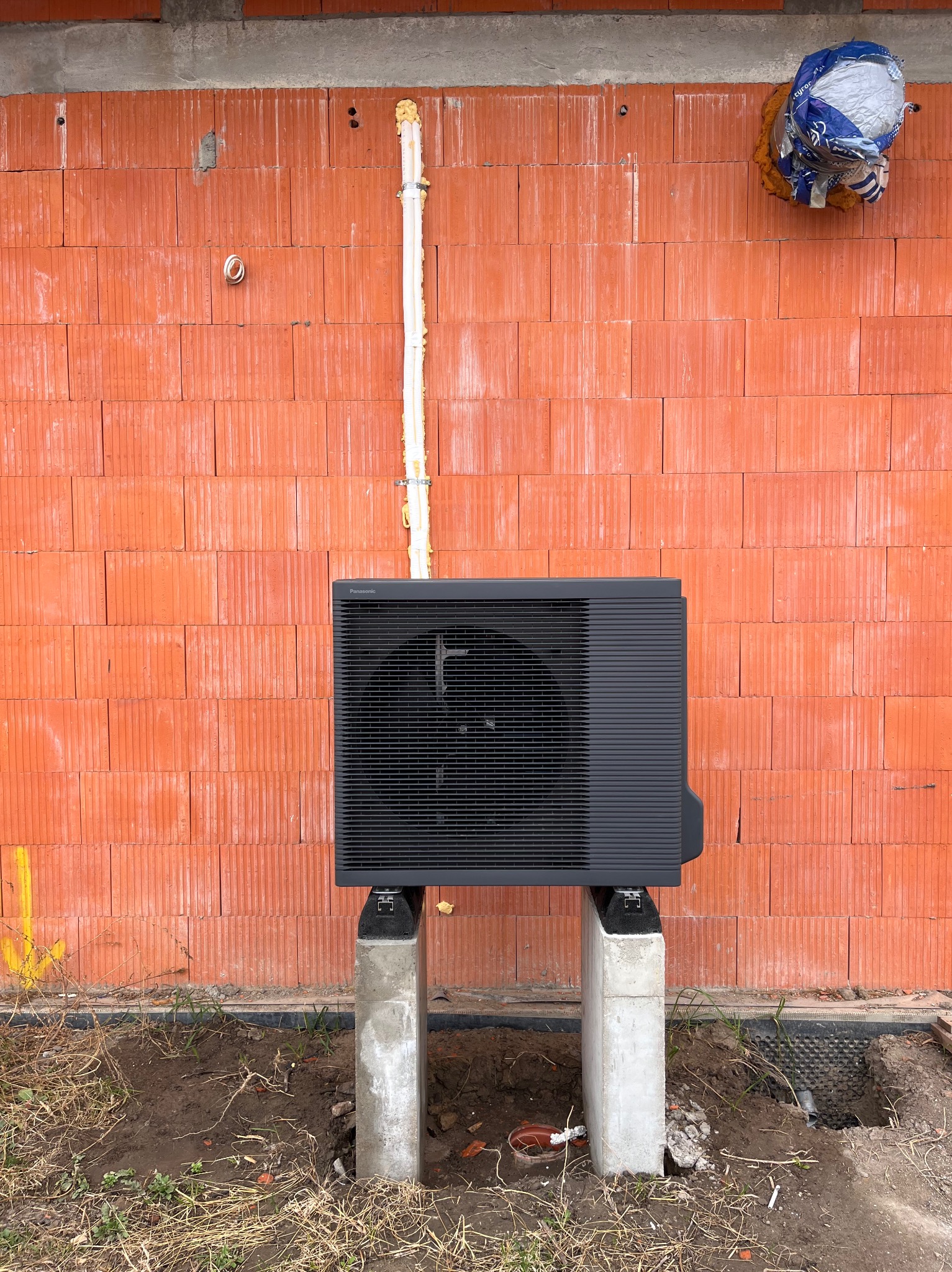 Czarna pompa ciepła Panasonic zamontowana na betonowych wspornikach przed ceglaną ścianą budynku w trakcie budowy. Widoczne rury i izolacja.