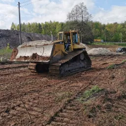 Żółty buldożer Caterpillar na placu budowy z ziemią na lemieszu, w tle hałda ziemi, drzewa i droga.
