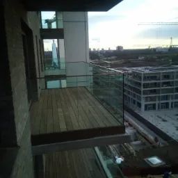 Nowoczesny balkon z drewnianą podłogą i szklaną balustradą, widok na miasto z dźwigami budowlanymi w tle.