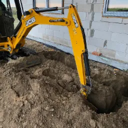 Żółta mini-koparka JCB w Toruniu podczas prac ziemnych, głęboki wykop przy fundamentach budynku z białego bloczka.