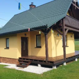 Żółty dom z ciemnobrązowymi elementami drewnianymi, zielonym dachem i małym balkonem, otoczony trawnikiem i białym żwirem, z praniem suszącym się na sznurku.