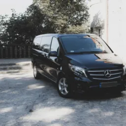 Czarny Mercedes Vito zaparkowany na brukowanym placu przed jasnym budynkiem z wieżyczką, otoczony zielenią, w słoneczny dzień.