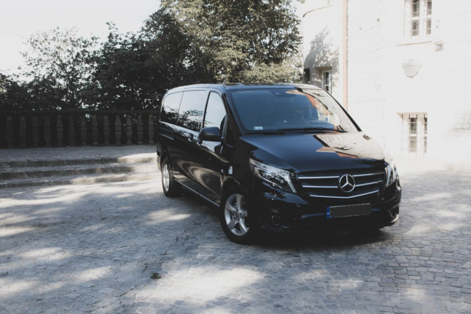 Czarny Mercedes Vito zaparkowany na brukowanym placu przed jasnym budynkiem z wieżyczką, otoczony zielenią, w słoneczny dzień.
