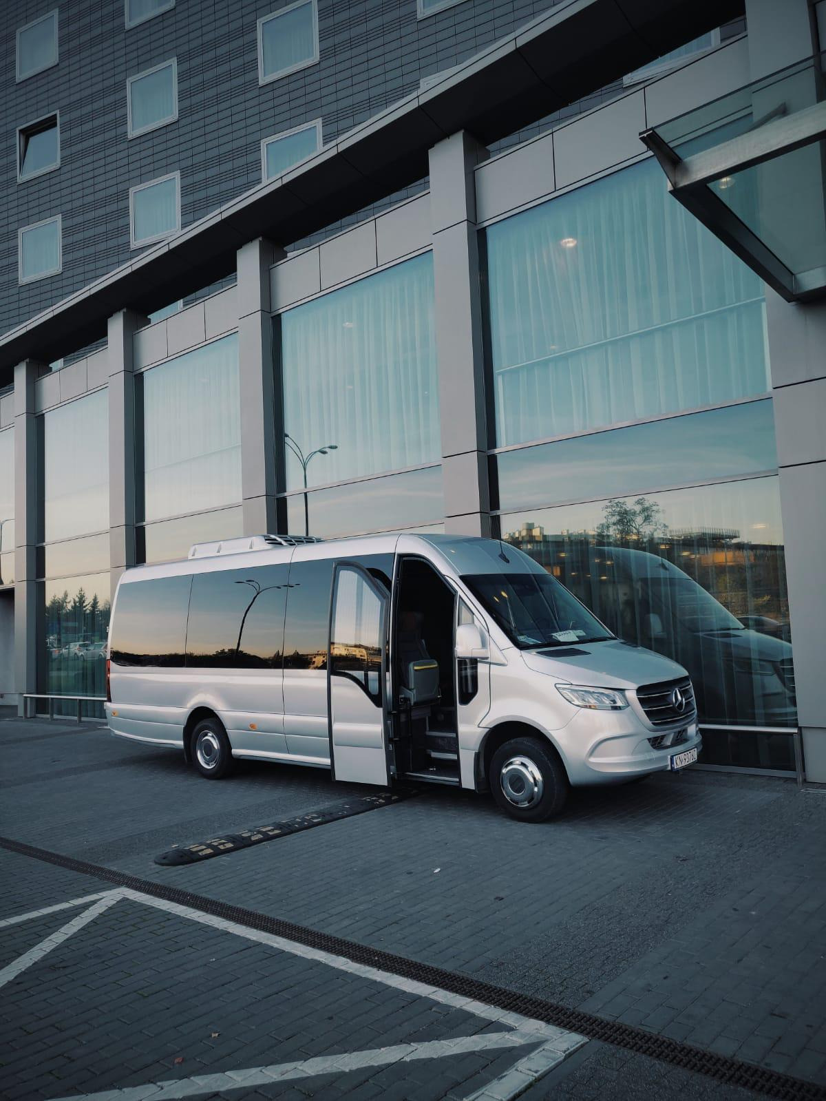 Srebrny bus Mercedes-Benz Sprinter z otwartymi drzwiami pasażera, zaparkowany przed nowoczesnym budynkiem z dużymi oknami, w których odbija się zachodzące słońce.