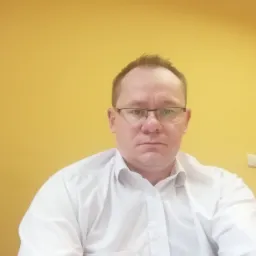 Portret mężczyzny w okularach i białej koszuli na tle żółtej ściany.