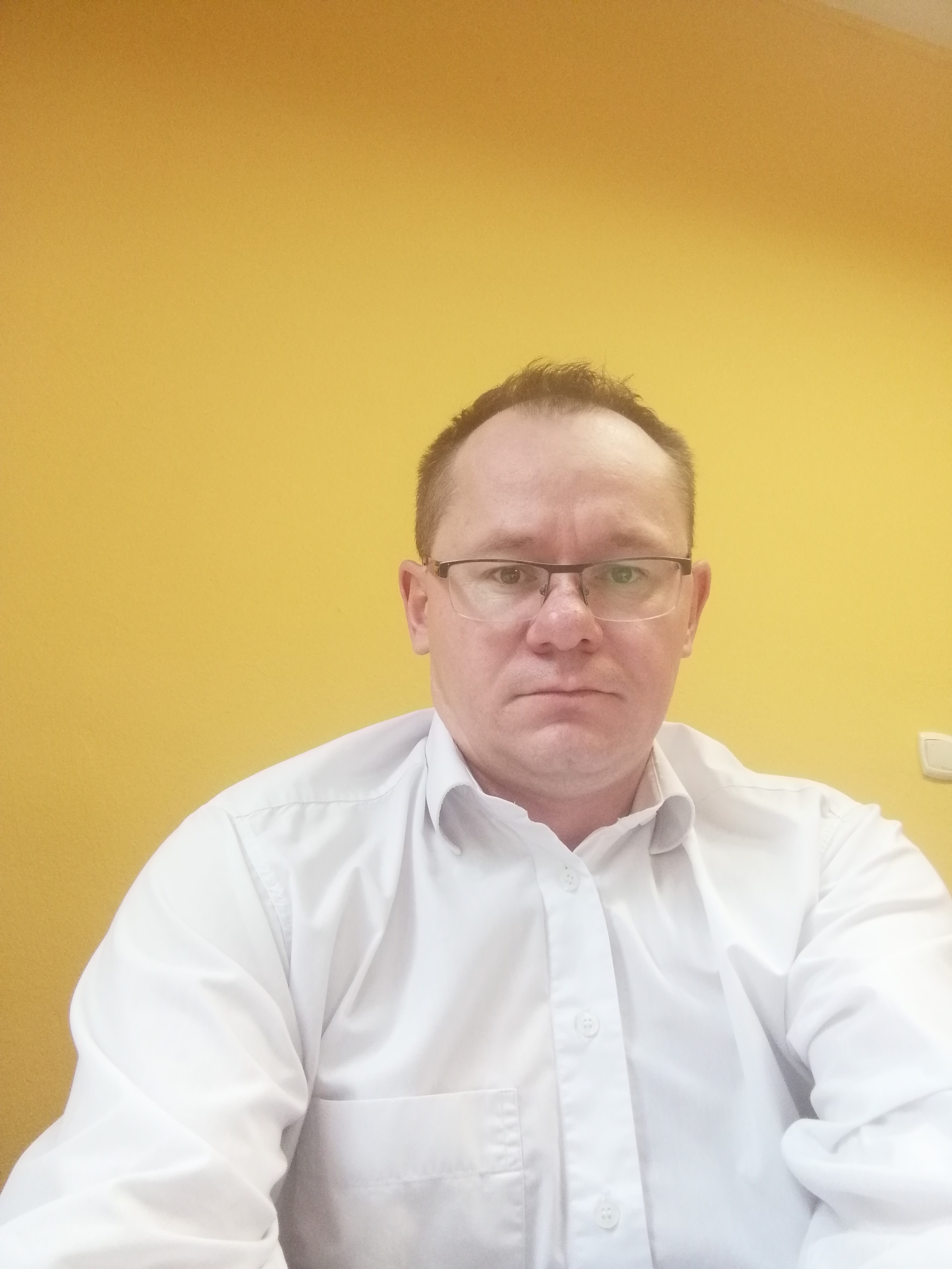 Portret mężczyzny w okularach i białej koszuli na tle żółtej ściany.
