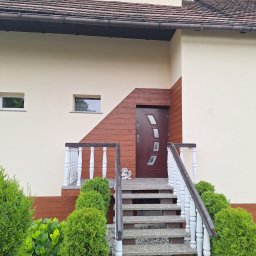 Pro-line - Wejście do domu z brązowymi drzwiami i schodami z szarego betonu z białą balustradą, otoczone zielenią i czerwonymi kwiatami, z fragmentem elewacji w kolorze kremowym i brązowym.