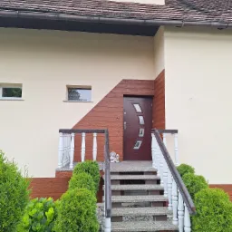 Wejście do domu z brązowymi drzwiami i schodami z szarego betonu z białą balustradą, otoczone zielenią i czerwonymi kwiatami, z fragmentem elewacji w kolorze kremowym i brązowym.