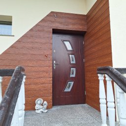 Pro-line - Wejście do domu z brązowymi drzwiami z szybami, obłożone drewnopodobną elewacją, z figurką aniołka na schodach.