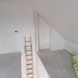 Poddasze w trakcie remontu: ocieplone ściany, sufit i podłoga, aluminiowa drabina oparta o ścianę, widoczne puszki elektryczne i worki z materiałem izolacyjnym.