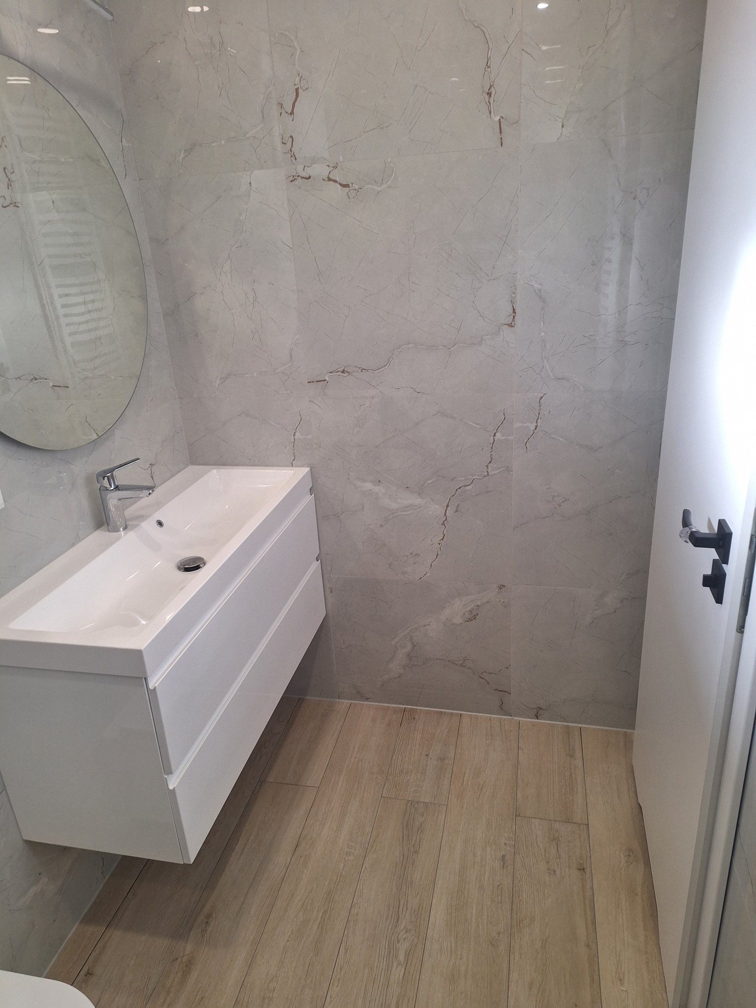 Nowoczesna łazienka z umywalką nablatową na białej szafce, okrągłe lustro i marmurowe płytki ścienne.