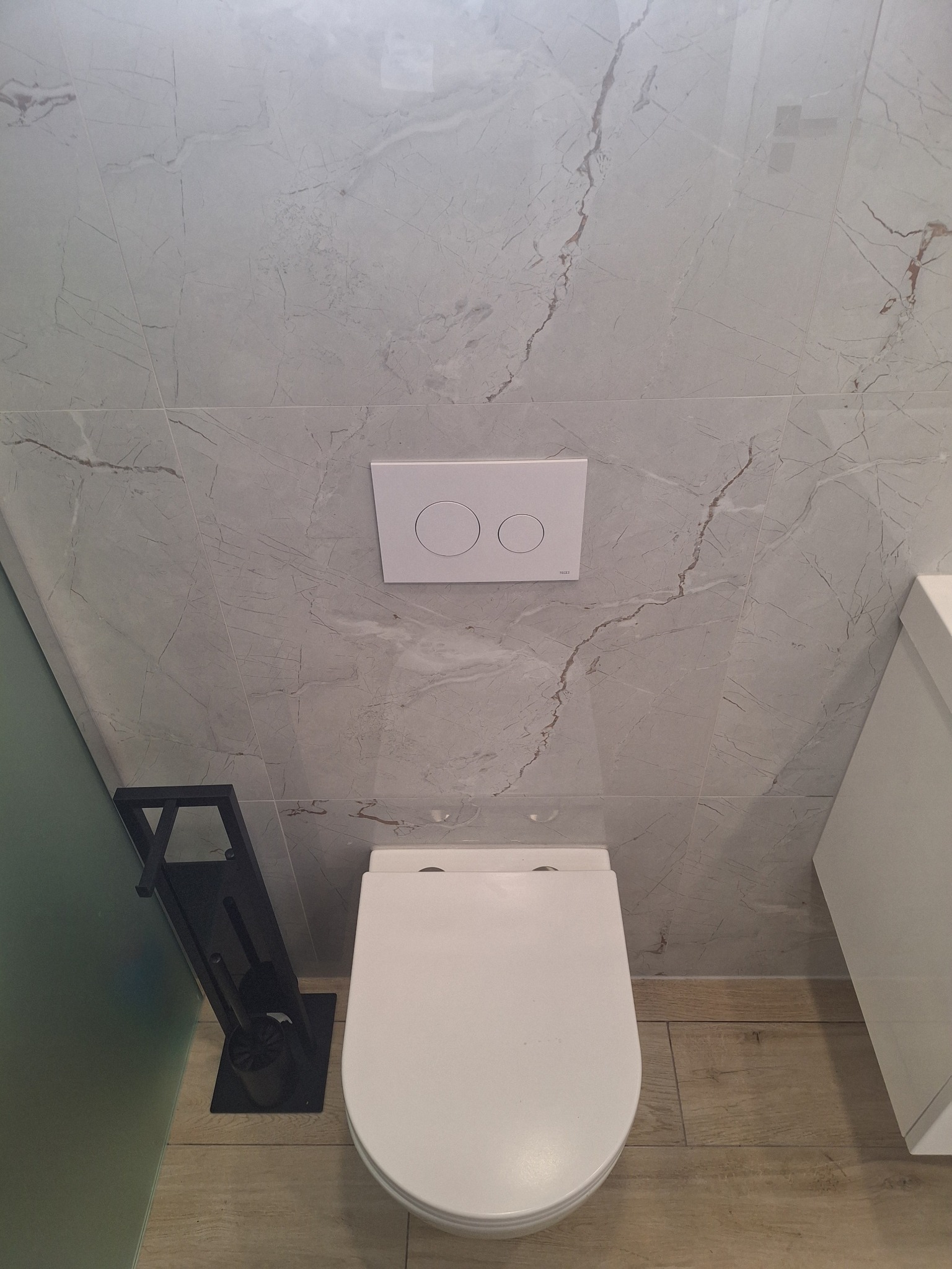 Wnętrze łazienki z wiszącą toaletą, przyciskiem spłukującym na ścianie z płytek imitujących marmur, oraz ciemnym stojakiem na szczotkę toaletową.