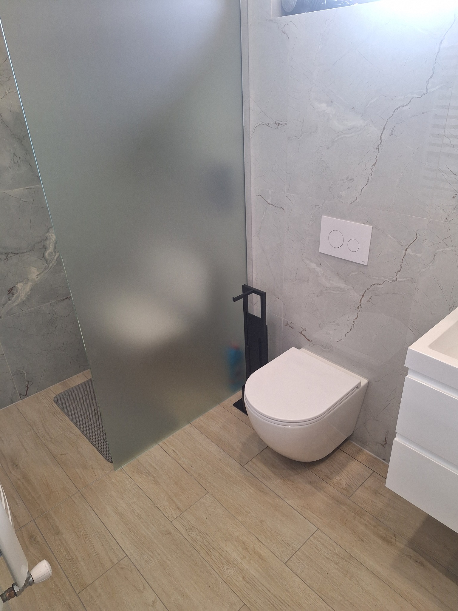 Nowoczesna łazienka z podwieszaną toaletą, szklaną kabiną prysznicową i marmurowymi płytkami na ścianach, oświetlona jasnym światłem.