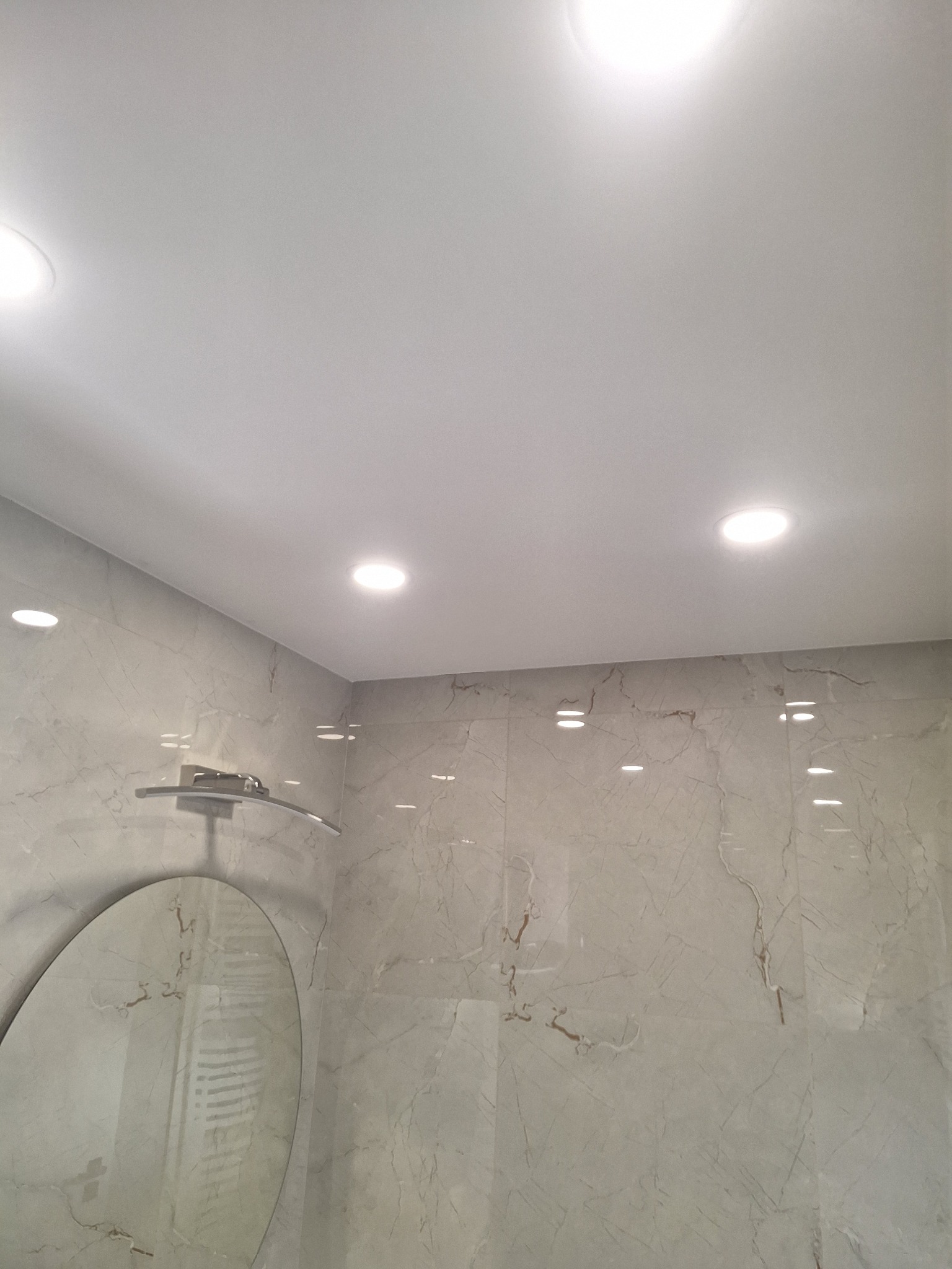 Wykończona łazienka z marmurowymi płytkami na ścianach, okrągłym lustrem i oświetleniem punktowym w suficie.