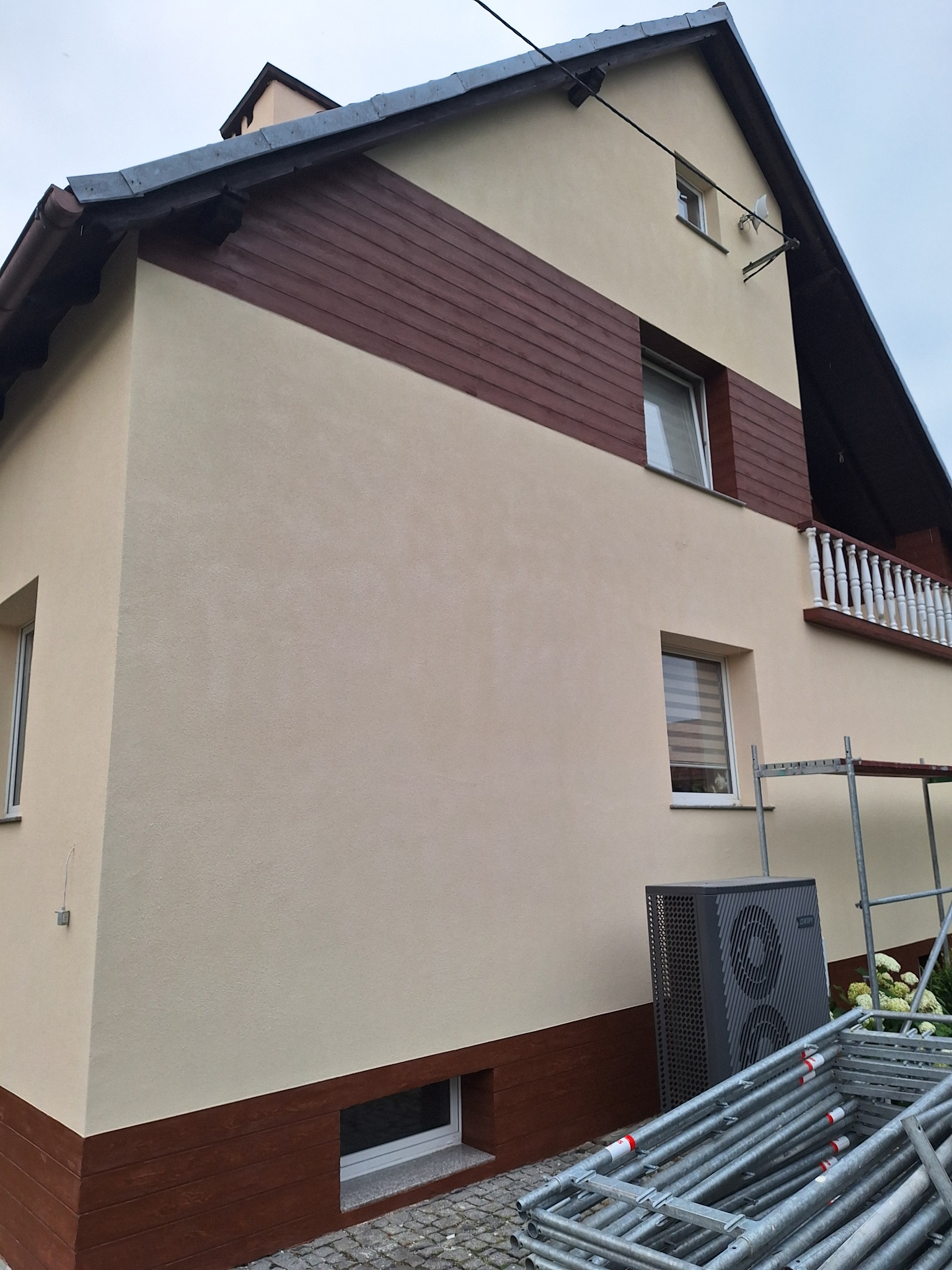 Dwukondygnacyjny dom z beżową elewacją i poziomym pasem brązowych desek, widoczny z boku, z ułożonymi rusztowaniami i pompą ciepła przy ścianie.