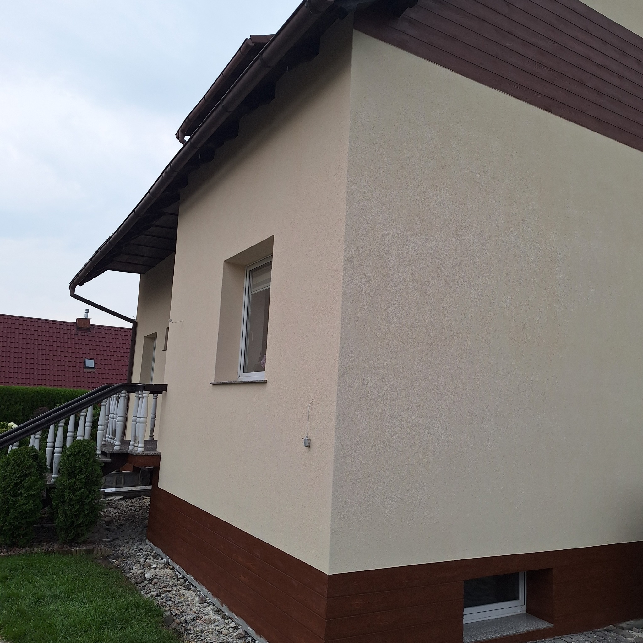 Jasna elewacja domu z brązową podbitką i podmurówką, widoczne okno piwniczne i fragment schodów wejściowych.