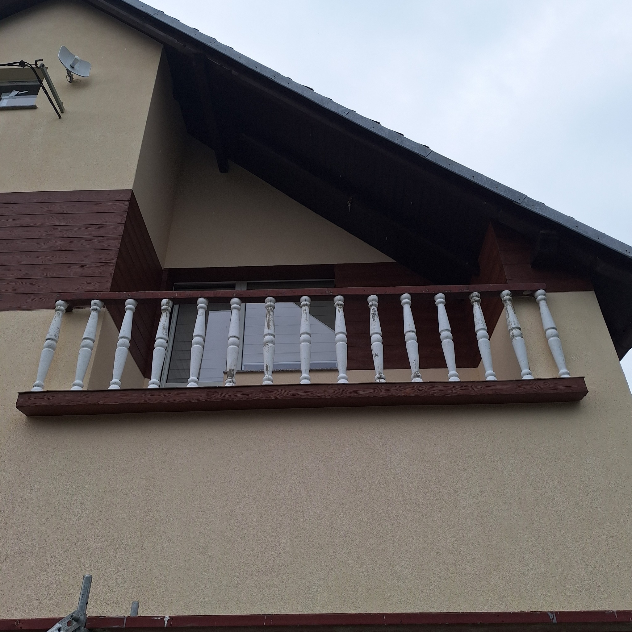 Fragment elewacji domu z balkonem o białych, toczonych balustradach i brązowym wykończeniu, widoczna antena satelitarna i fragment rusztowania.