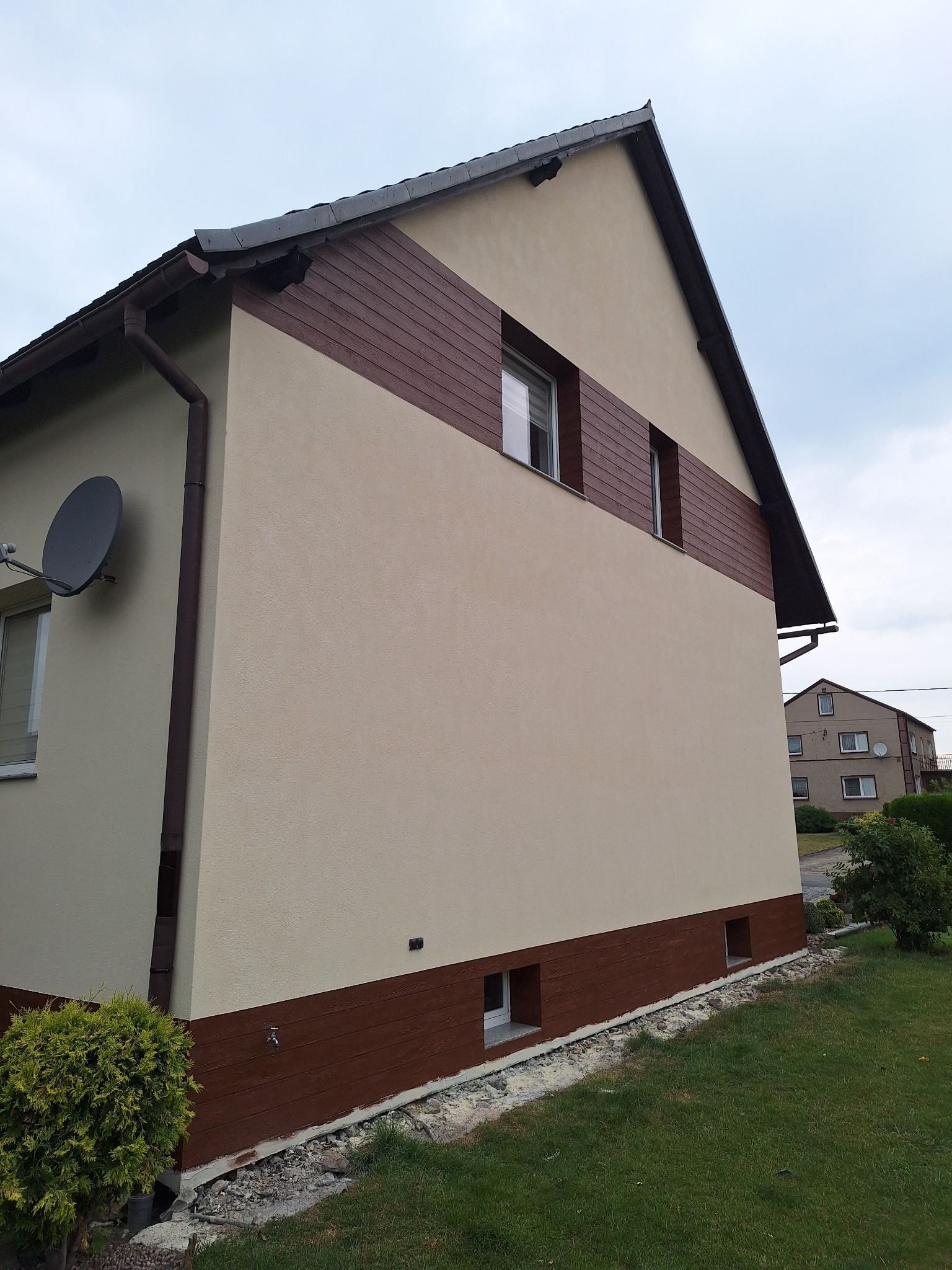 Wykończona elewacja domu jednorodzinnego z brązowym pasem imitującym drewno, widoczne okna i antena satelitarna, trawnik przed budynkiem.