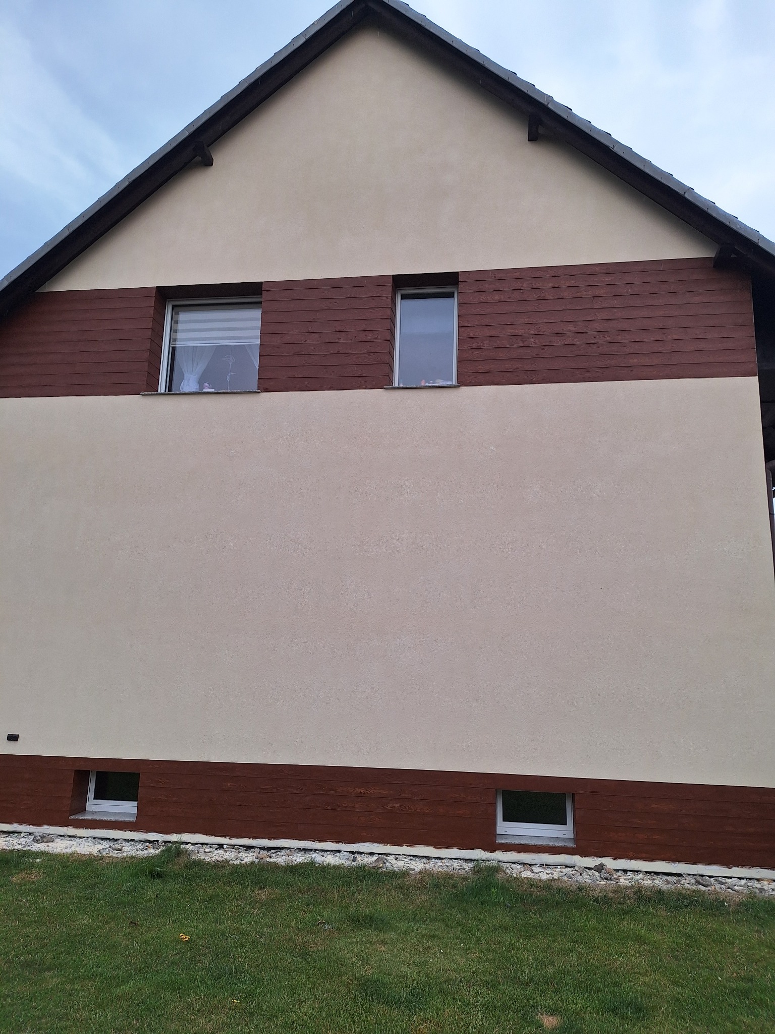 Dwukondygnacyjny dom z beżową elewacją i poziomymi panelami imitującymi drewno, widoczne okna z roletami w górnej części i małe okienka przy gruncie, trawnik z przodu.