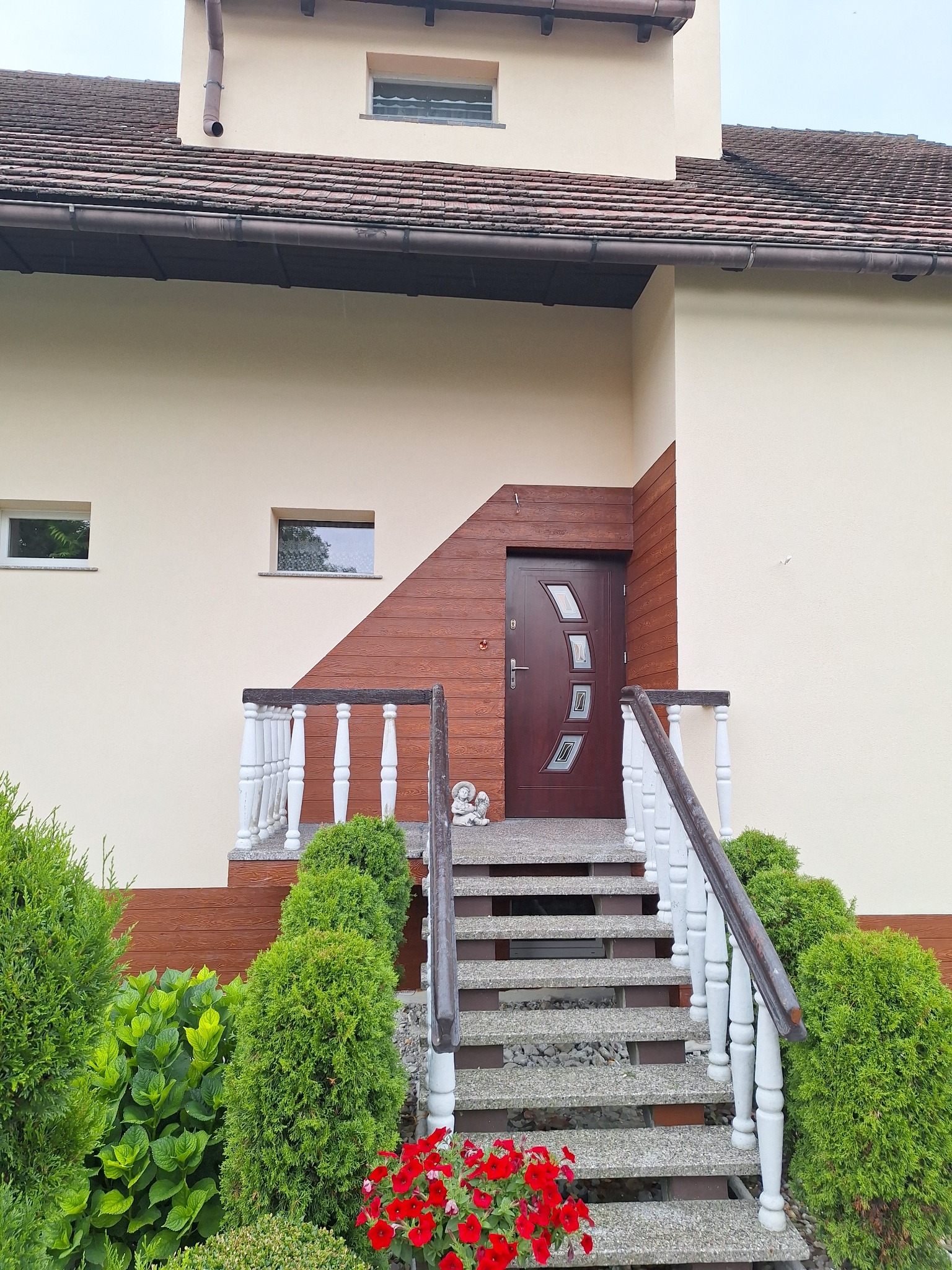 Wejście do domu z brązowymi drzwiami i schodami z szarego betonu z białą balustradą, otoczone zielenią i czerwonymi kwiatami, z fragmentem elewacji w kolorze kremowym i brązowym.