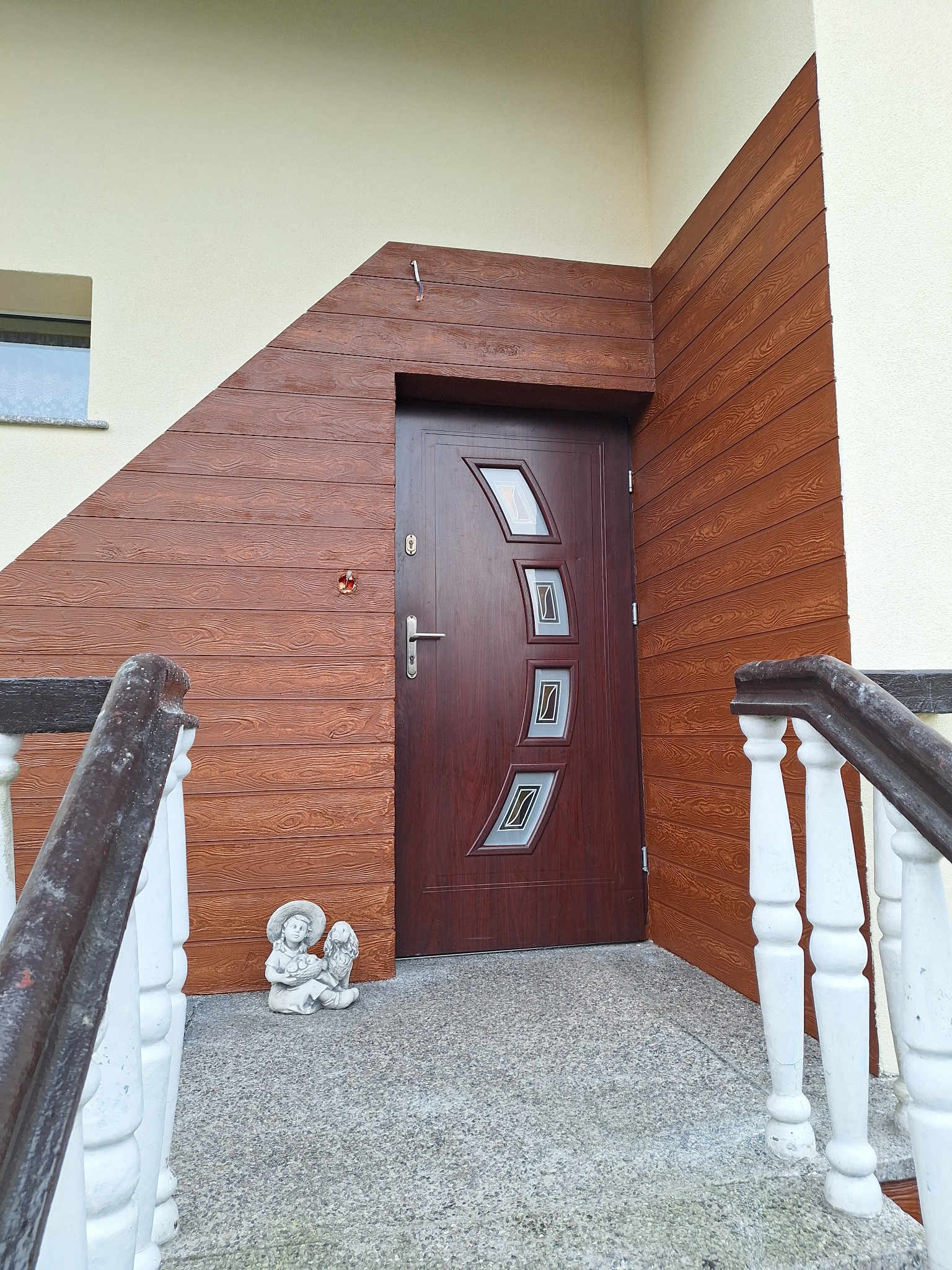 Wejście do domu z brązowymi drzwiami z szybami, obłożone drewnopodobną elewacją, z figurką aniołka na schodach.