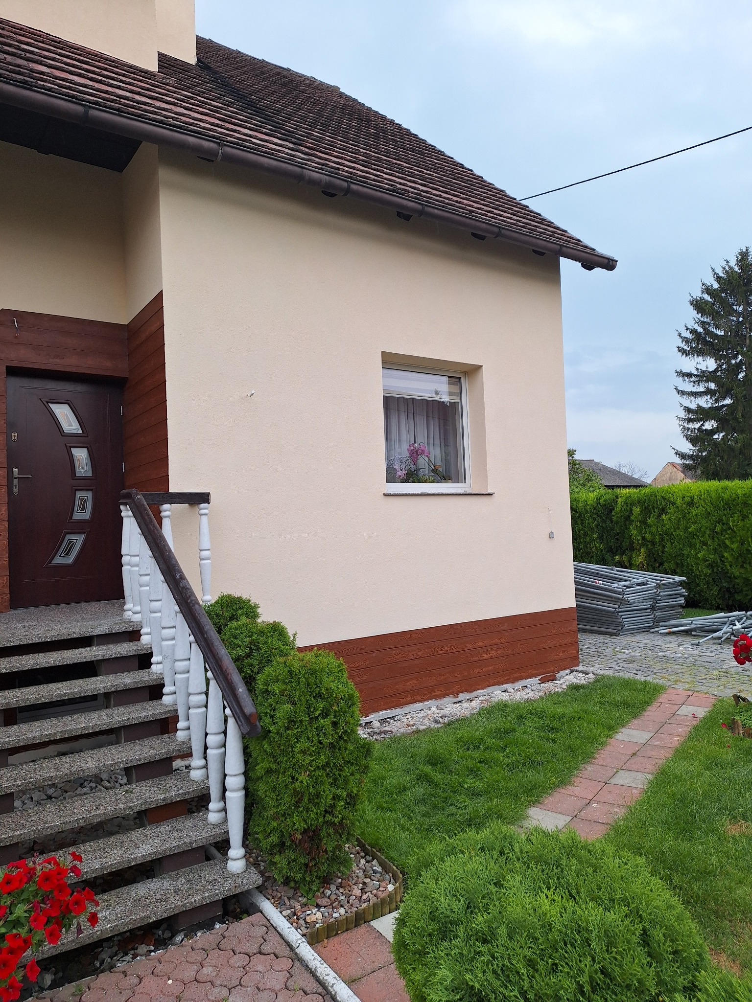 Dom jednorodzinny z beżową elewacją, brązową podbitką i schodami wejściowymi z białymi balustradami, w tle widoczne rusztowania.