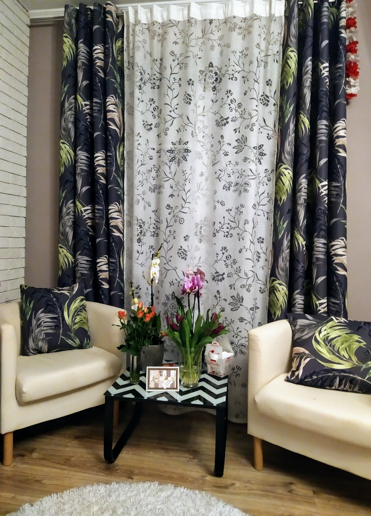 Dwa kremowe fotele z poduszkami w liście, rozdzielone stolikiem z kwiatami, na tle zasłon w botaniczne wzory: ciemnej z liśćmi i jasnej z kwiecistymi ornamentami.