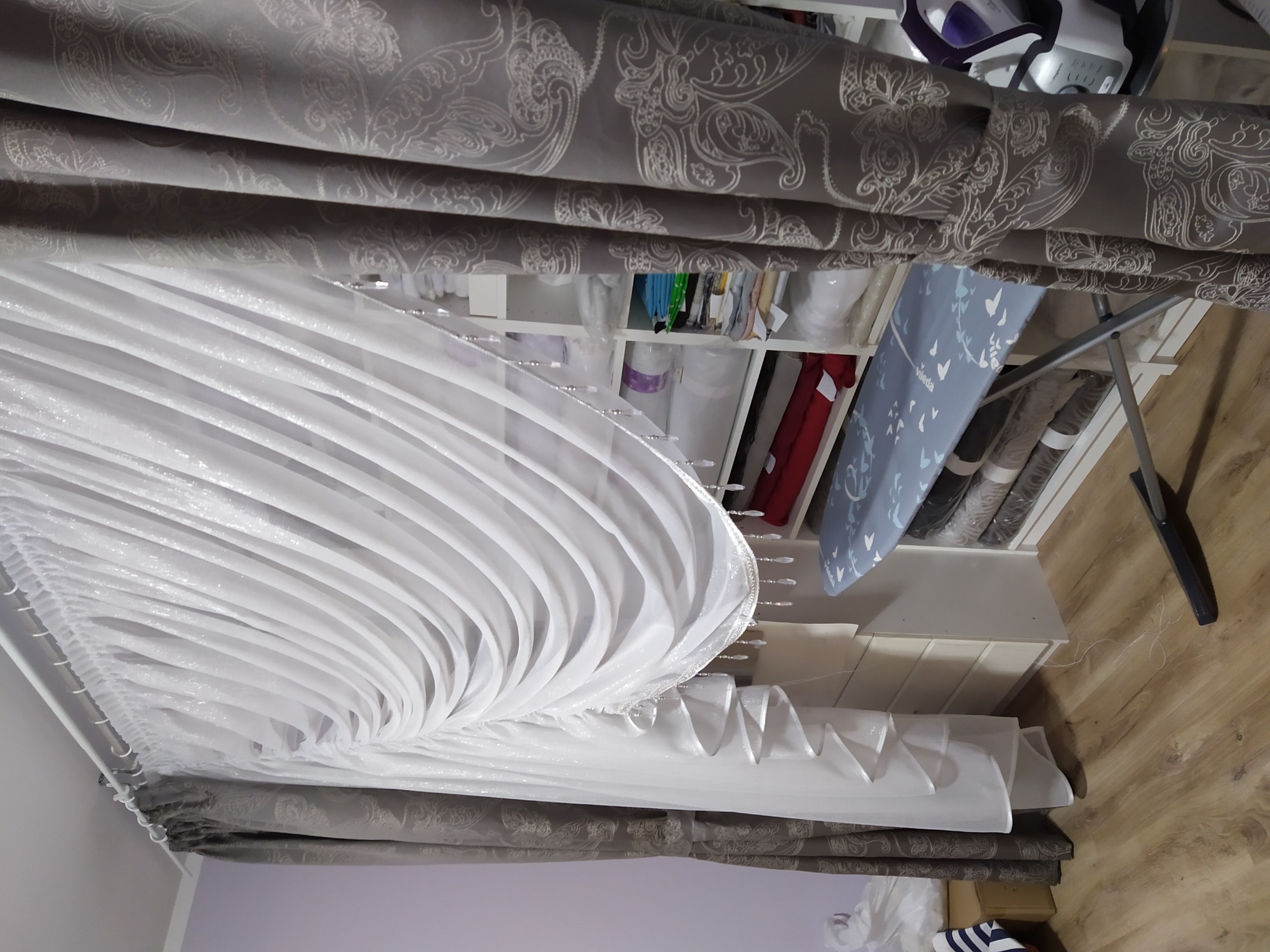 Biała, plisowana firanka z ozdobnymi koralikami, przygotowana do montażu w Bieruniu, z widocznym fragmentem zasłony w odcieniach szarości.