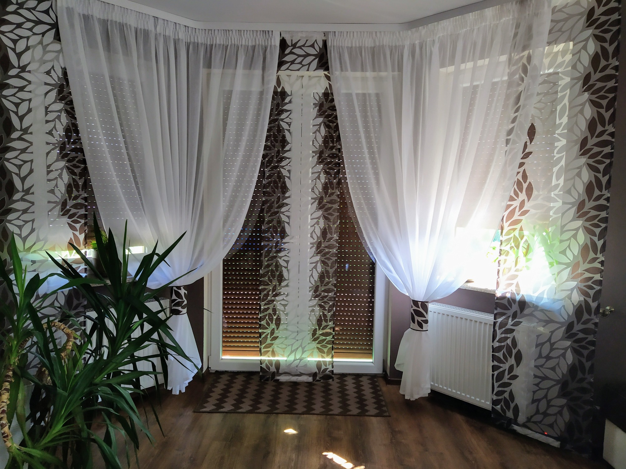 Jasne, zwiewne zasłony z motywem liści na oknie z roletami, w tle grzejnik i roślina doniczkowa.