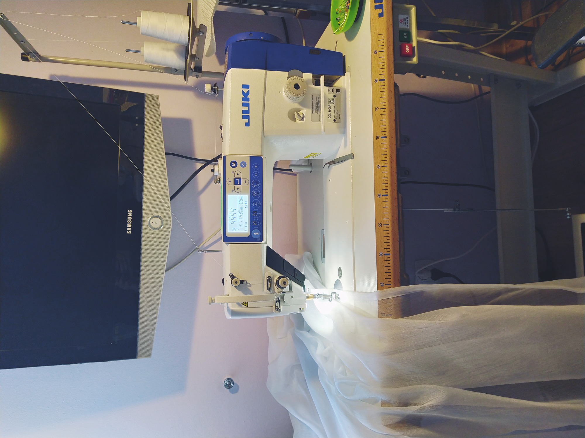 Profesjonalna maszyna do szycia JUKI z elektronicznym wyświetlaczem, szyjąca delikatną, białą tkaninę, widoczna miarka drewniana, tło stanowią elementy wyposażenia pracowni.
