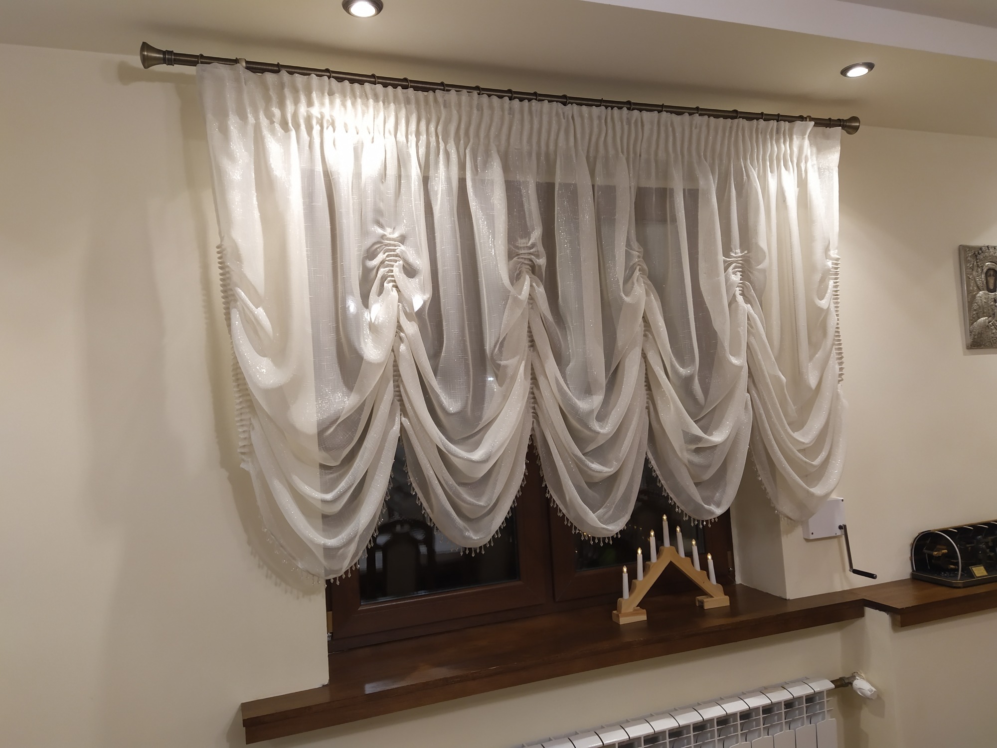 Elegancka, drapowana, biała zasłona z połyskującą nitką i koralikami, zawieszona na ciemnym karniszu nad oknem z drewnianą ramą, na parapecie drewniany świecznik adwentowy i korbka do rolet.