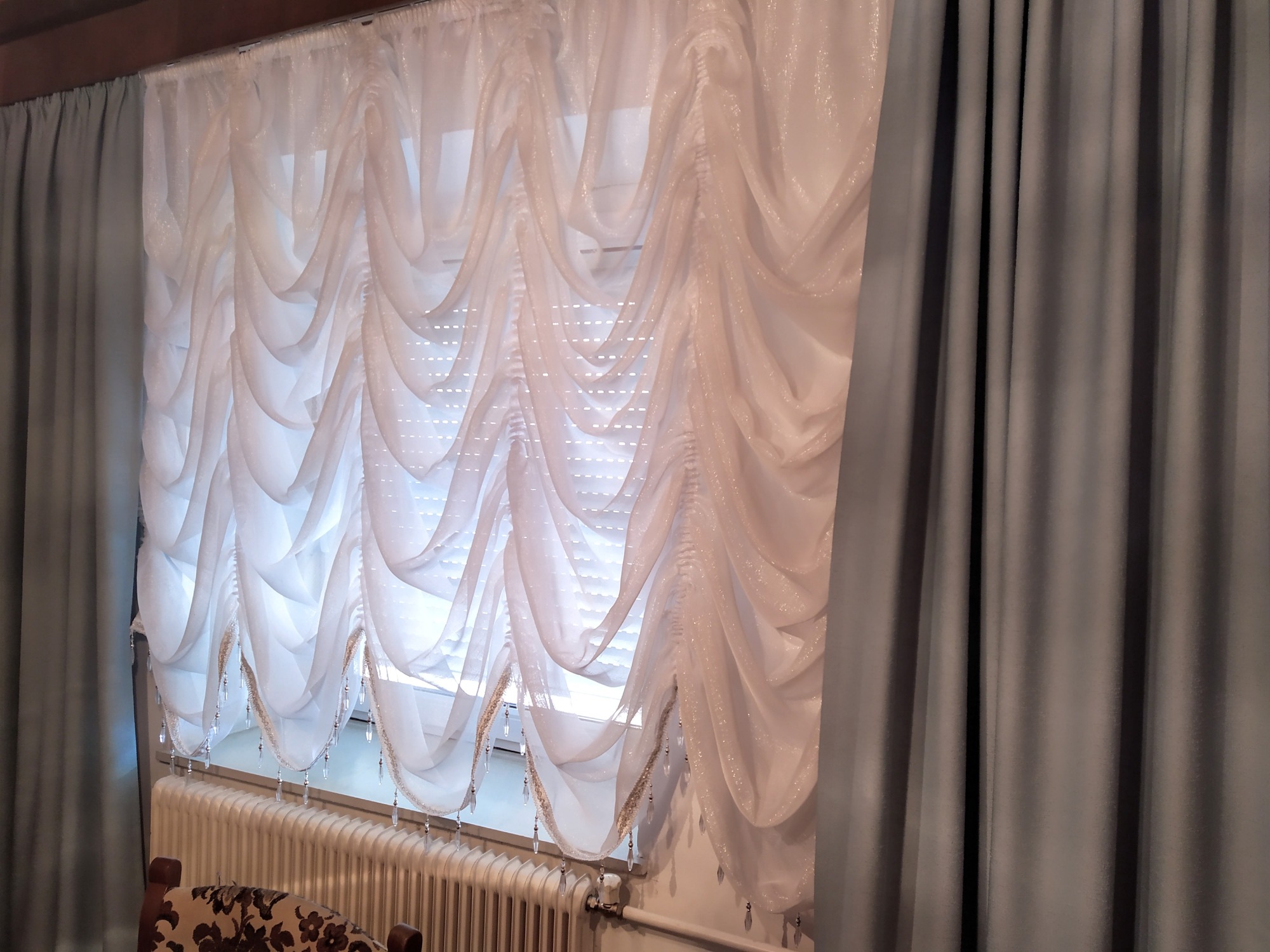 Okno z białymi, zwiewnymi firanami z kryształkami, zasłonięte po bokach szarymi zasłonami.