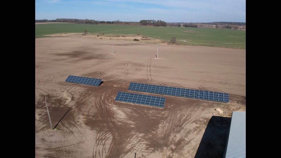 Widok z lotu ptaka na rozłożone panele słoneczne na przygotowanym polu, widoczne ślady opon i słupy energetyczne w tle, częściowo zadaszony budynek w prawym dolnym rogu.