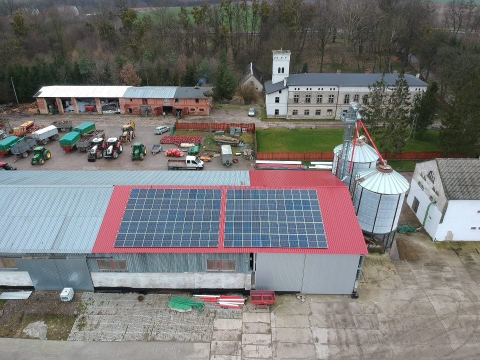 Widok z góry na budynek gospodarczy z czerwonym dachem, na którym zamontowano dwa panele słoneczne; w tle zabudowania gospodarcze, silosy i dwór z wieżą.