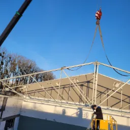 Pracownik na podnośniku koszowym, montuje metalową konstrukcję ogrodzenia, podnoszoną przez dźwig na tle błękitnego nieba.