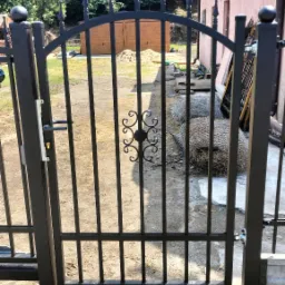 Czarna, metalowa brama z ozdobnym elementem pośrodku, widok na podwórze z piaskiem i materiałami budowlanymi, świeżo zamontowana.