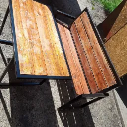 Stolik z drewnianym blatem i metalową ramą, widoczne ślady spawania, nierówna powierzchnia betonu w tle.