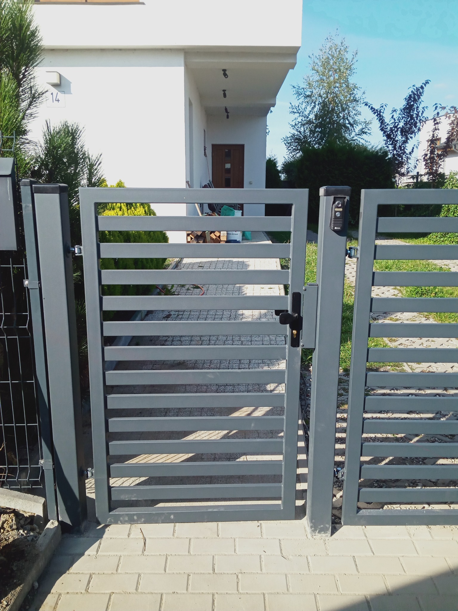 Nowoczesna, metalowa furtka z poziomymi szczeblami w kolorze grafitowym, prowadząca do domu z jasną elewacją i numerem 14, widoczna skrzynka na listy i elementy ogrodzenia.