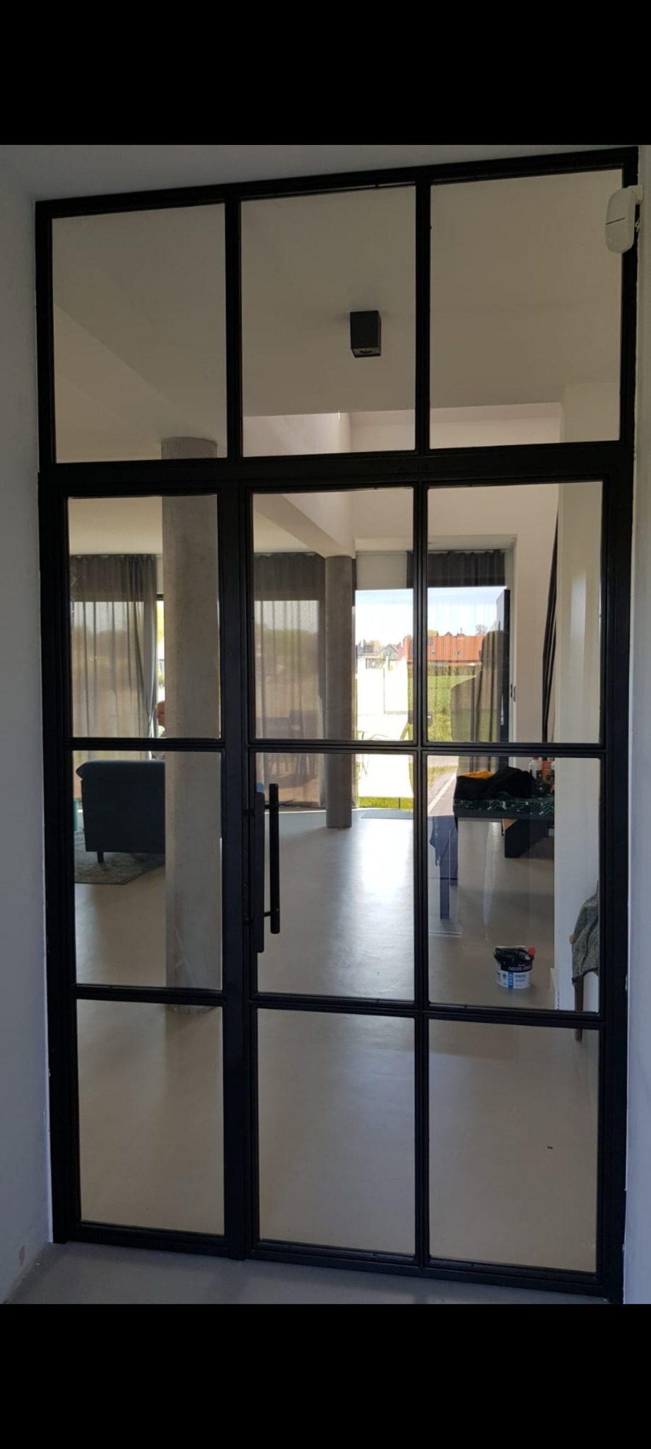 Wewnętrzne dwuskrzydłowe drzwi z czarną, metalową ramą i szklanymi panelami, widok na jasne wnętrze domu z betonowymi kolumnami i częściowo zasłoniętymi oknami.